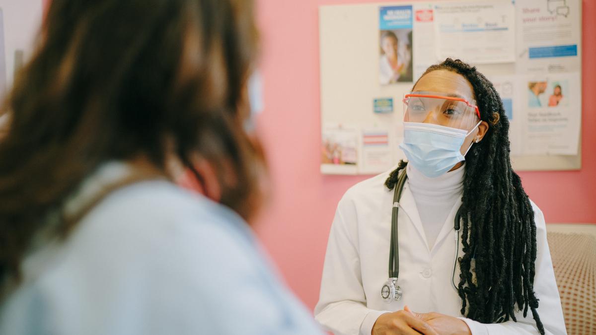 A medical staff member wearing a mask, sitting with a patient in an exam room