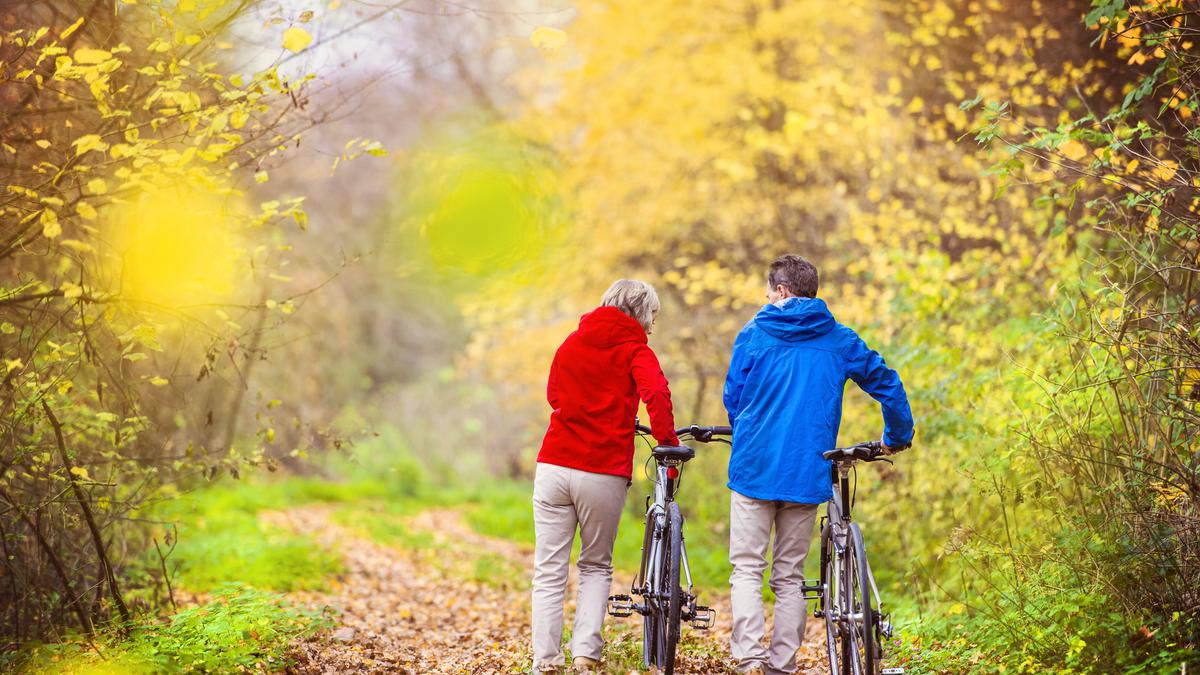 [image of two people in red and blue jackets and khakis pushing bikes on a green path in the woods.]