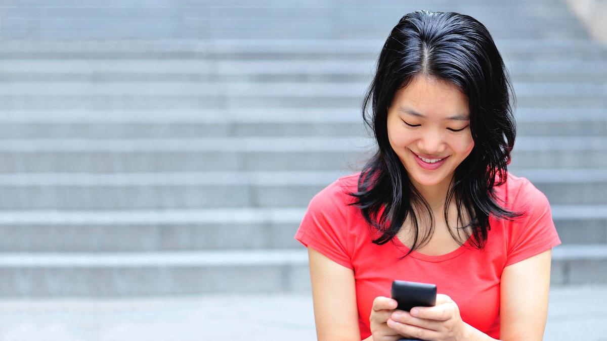 [image of a person in a red shirt with black hair looking down and smiling while on their phone. The background is a blurred image of gray stairs.]