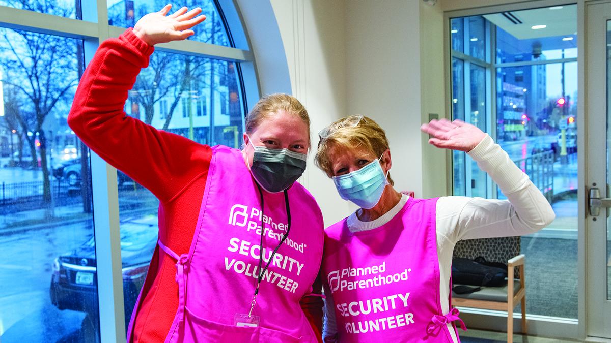 Two volunteers pose, waving at the camera. They both wear bright pink vests with the Planned Parenthood logo that say "Security Volunteer" on them.