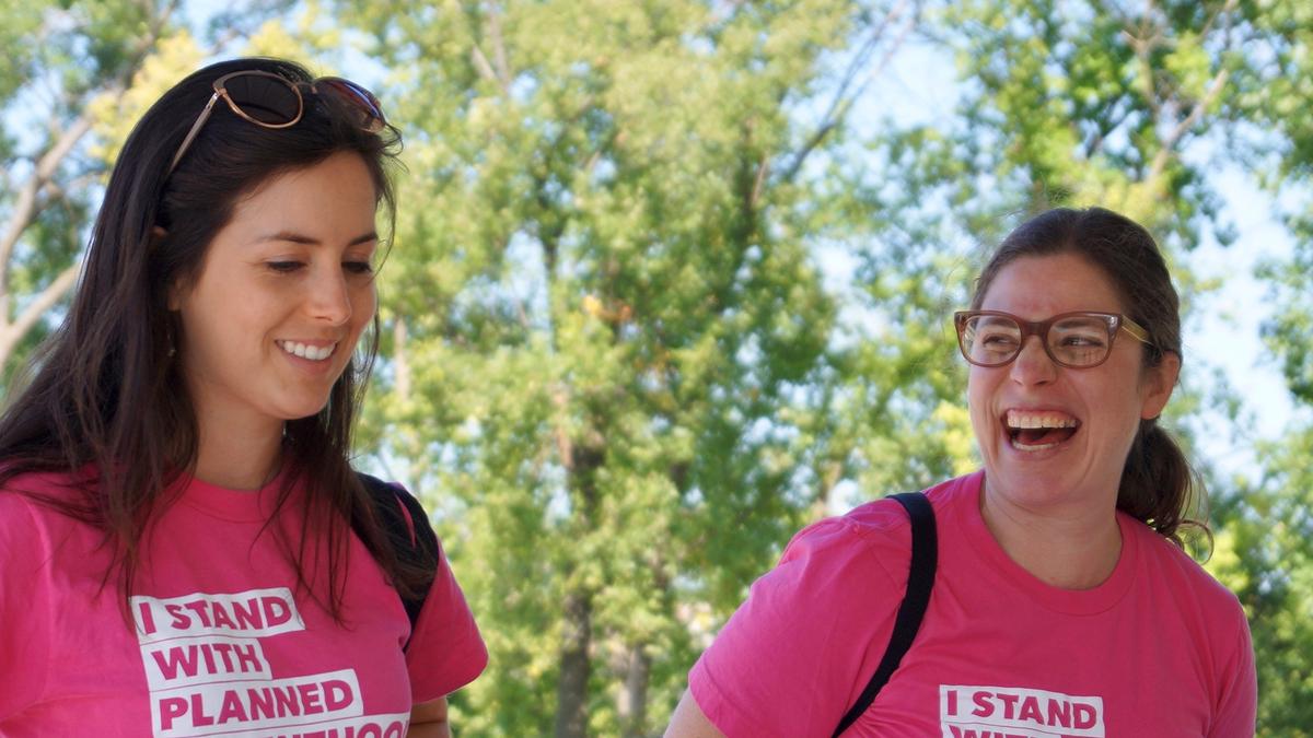 Two volunteers standing outside, smiling. Both wearing pink shirts that say "I stand with Planned Parenthood".