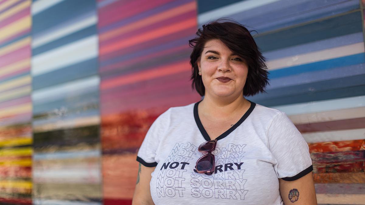 Planned Parenthood volunteer, Isa, smiles in front of a bright wall. She is wearing a shirt that says "Not Sorry."